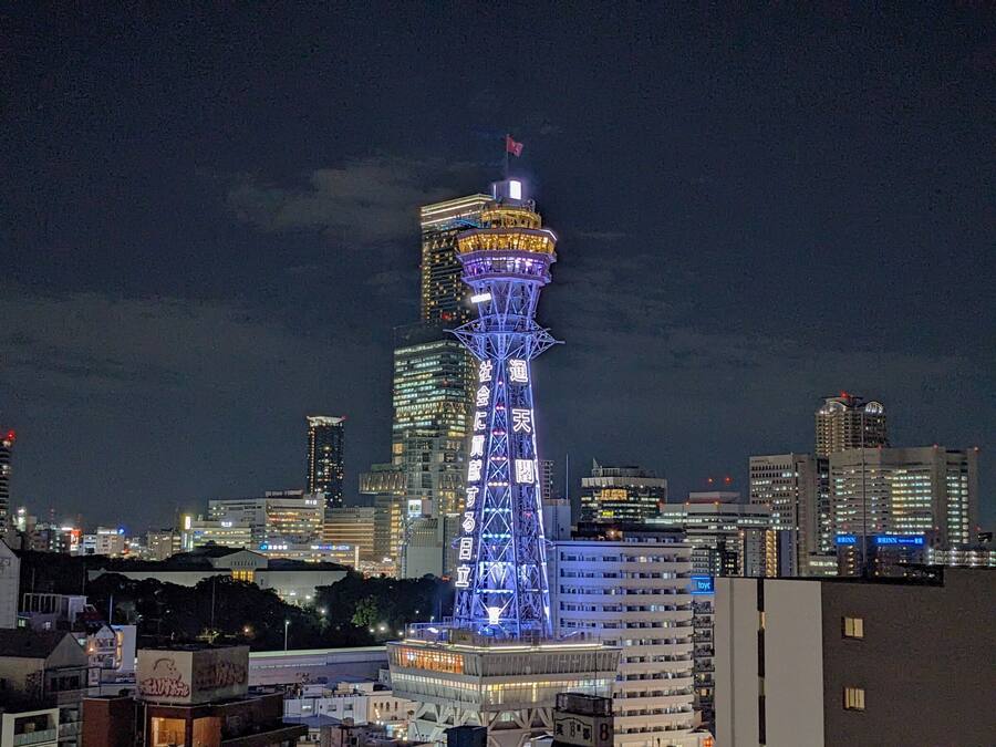 A tall, illuminated tower with a flag at the top, surrounded by a cityscape at night.