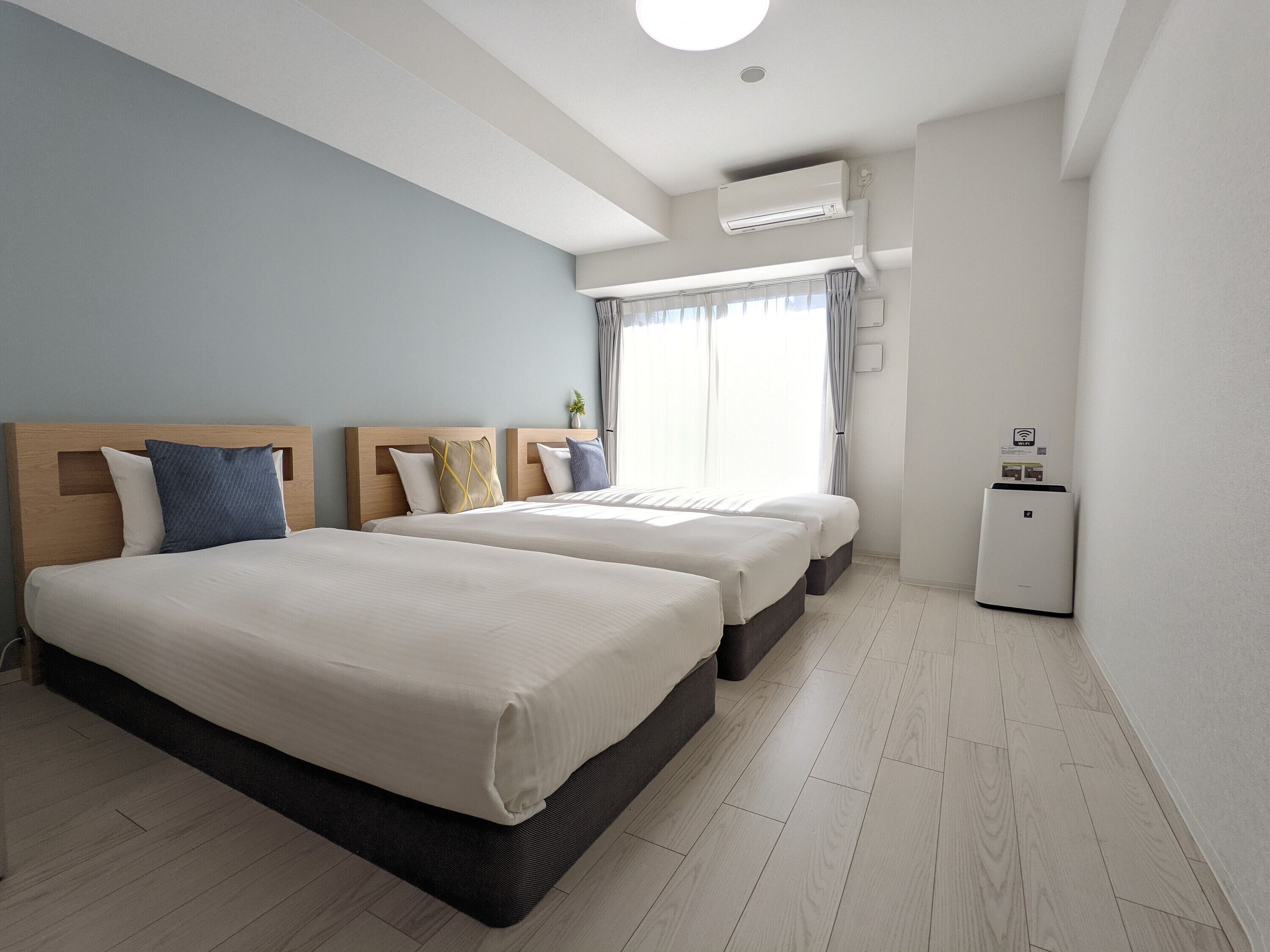 A hotel room with two beds, a small desk, a window with curtains, and an air conditioning unit.