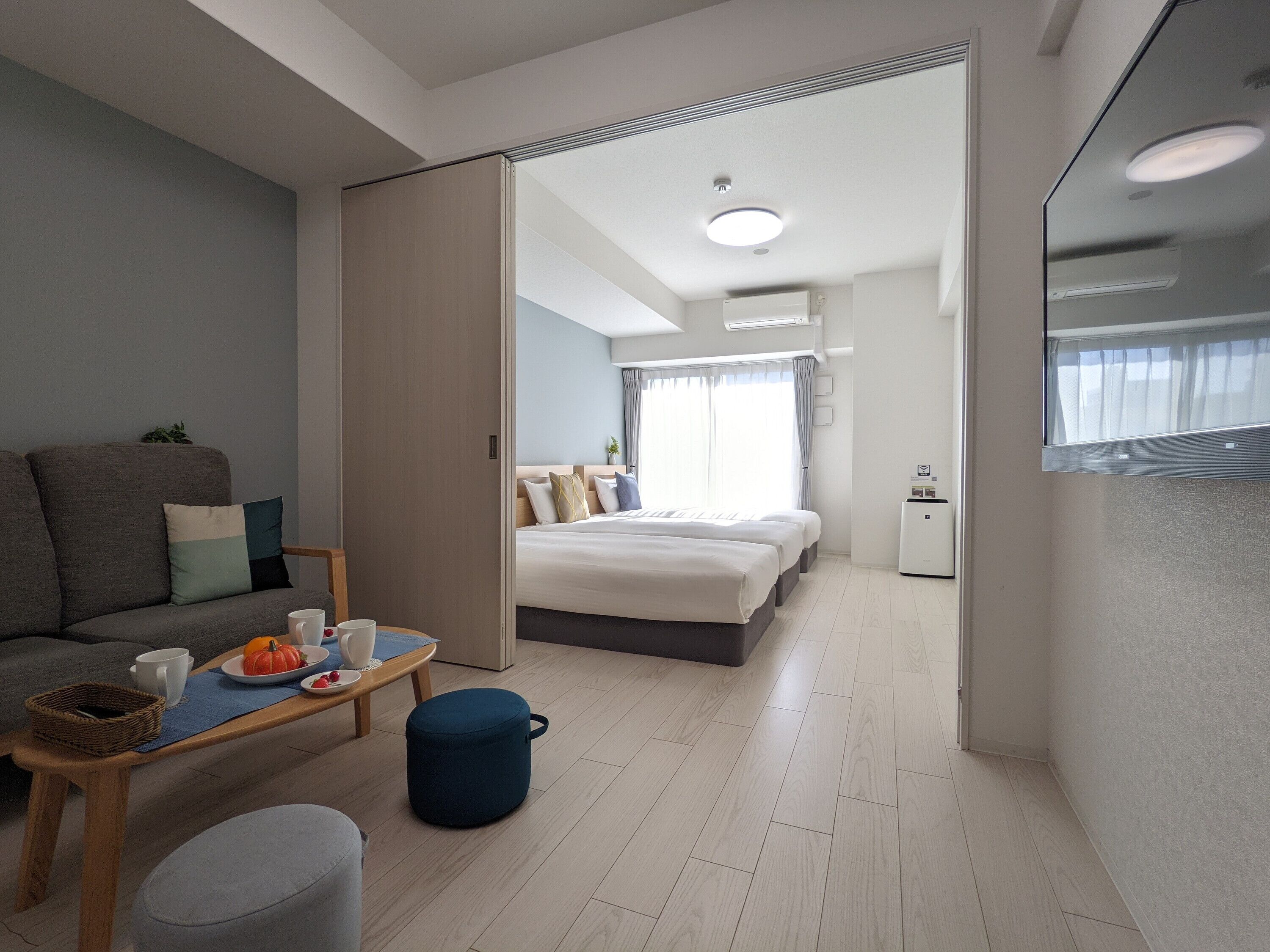 Quarto de hotel moderno com sofá, mesa de jantar com cesta, um banquinho azul e cama com cabeceira.