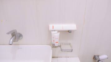 Casa de banho com lavatório branco, torneira, espelho e prateleira com produtos de higiene, incluindo escova de dentes e tubo de loção corporal.