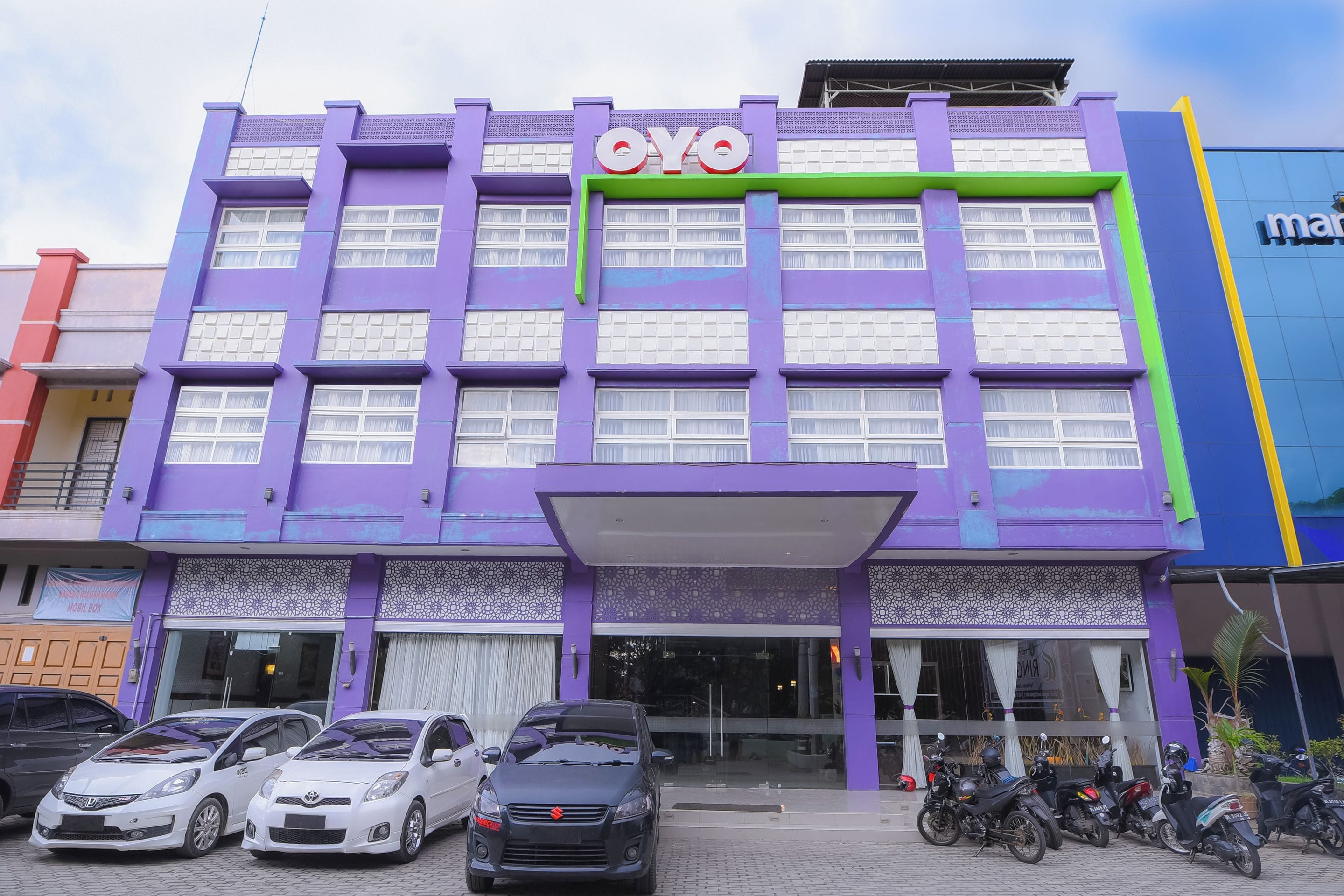 Ein mehrstöckiges Gebäude mit violett schimmernder Fassade, einem gut sichtbaren 'OYO'-Schild und mehreren parkenden Autos und Motorrädern davor.