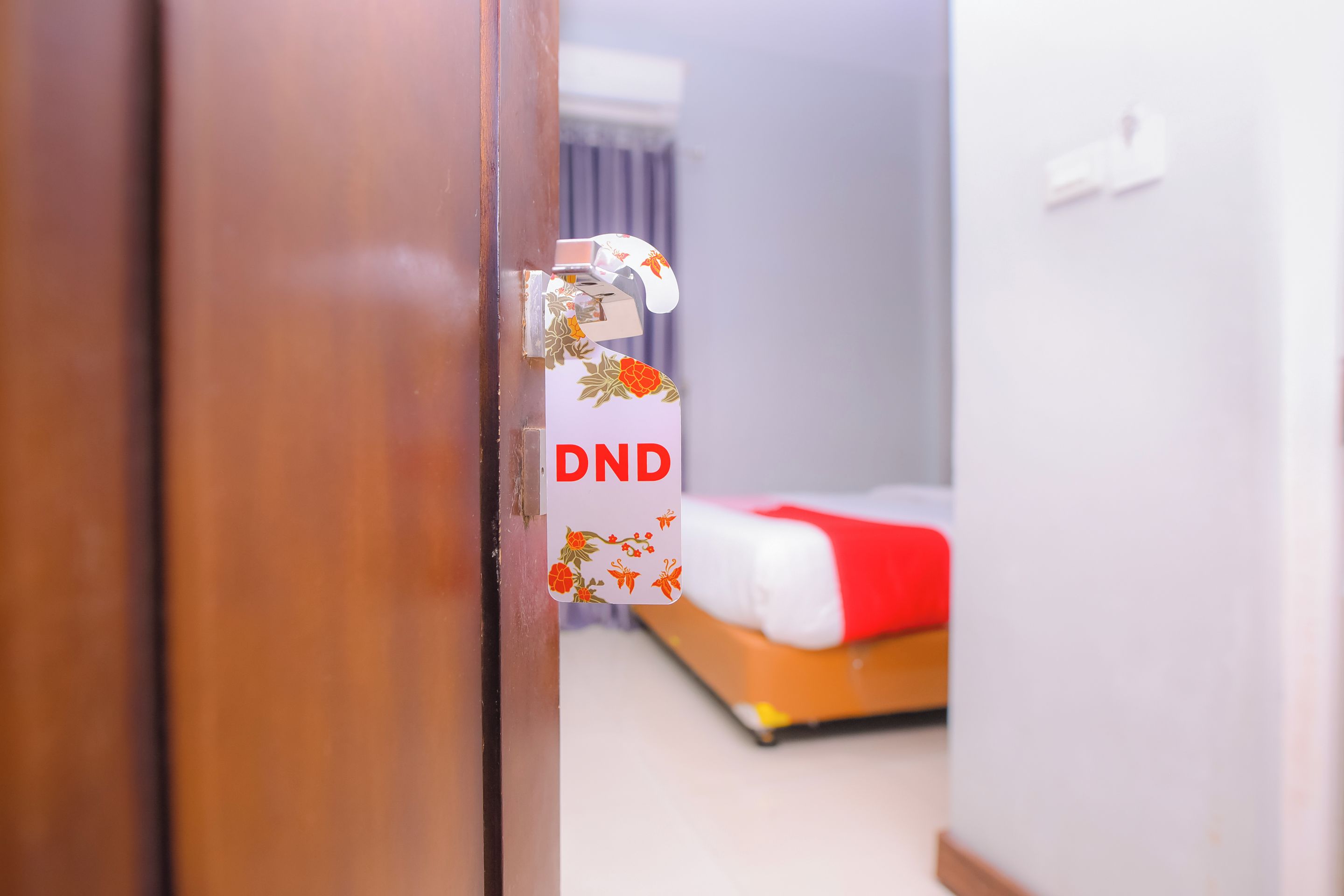 Eine Hotelzimmertür mit einem Bitte-nicht-stören-Schild, ein Bett mit roter und weißer Bettwäsche und ein Fenster mit Vorhängen.
