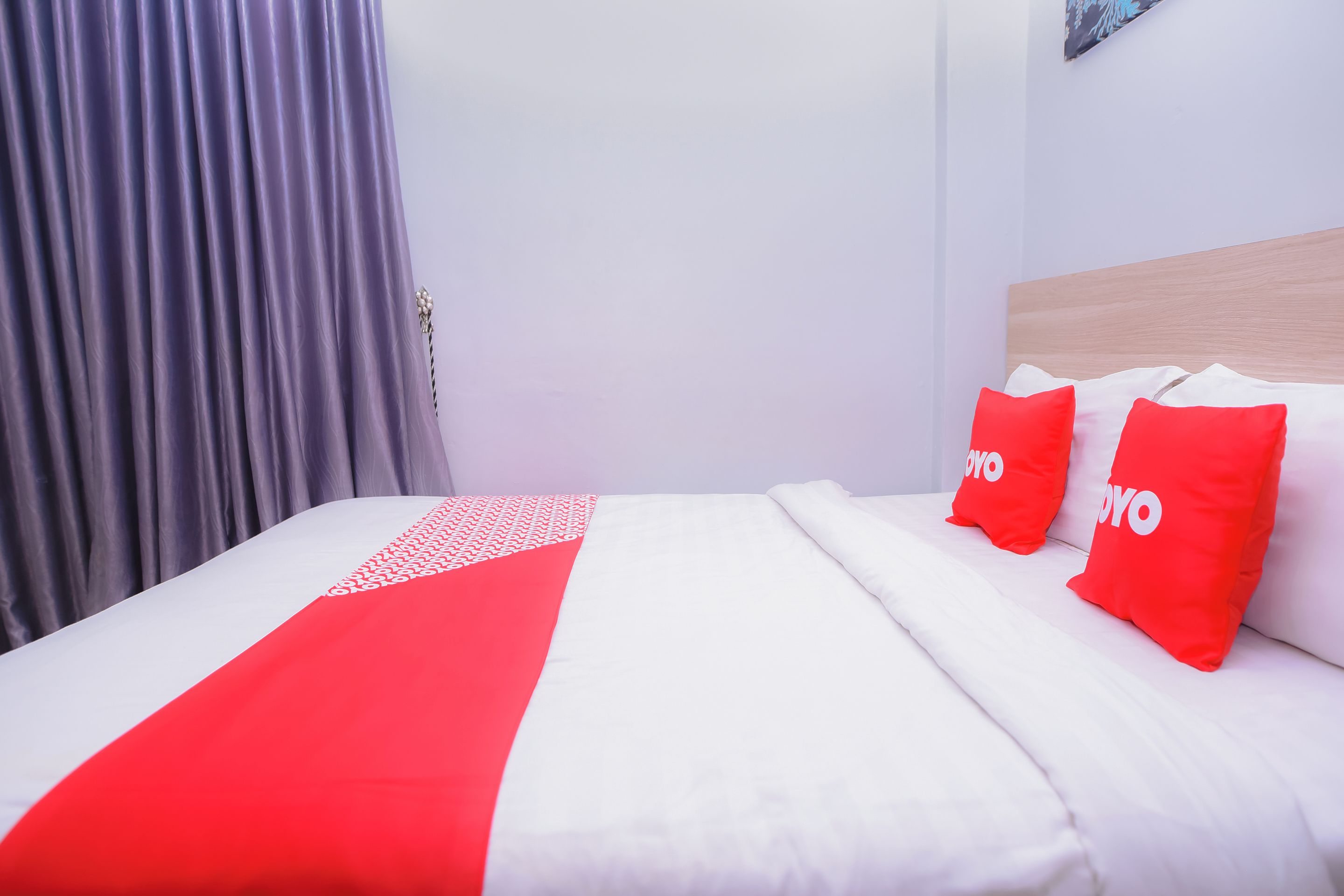 Ein Bett mit weißer und roter Bettwäsche, zwei rote Kissen mit dem Aufdruck 'YOYO' sowie ein grauer Vorhang und ein Holz-Kopfseitenverkleidung.
