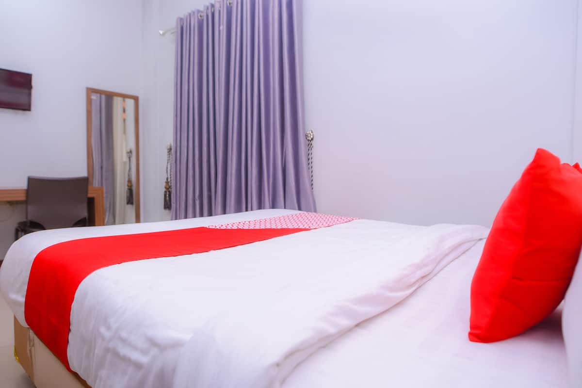 Ein ordentlich bezogenes Bett mit weißer und roter Bettwäsche, einem roten Kopfkissen und einem violetten Vorhang in einem schlichten Zimmer mit einem Holz-Kopfteil und einem kleinen Schreibtisch mit Stuhl.