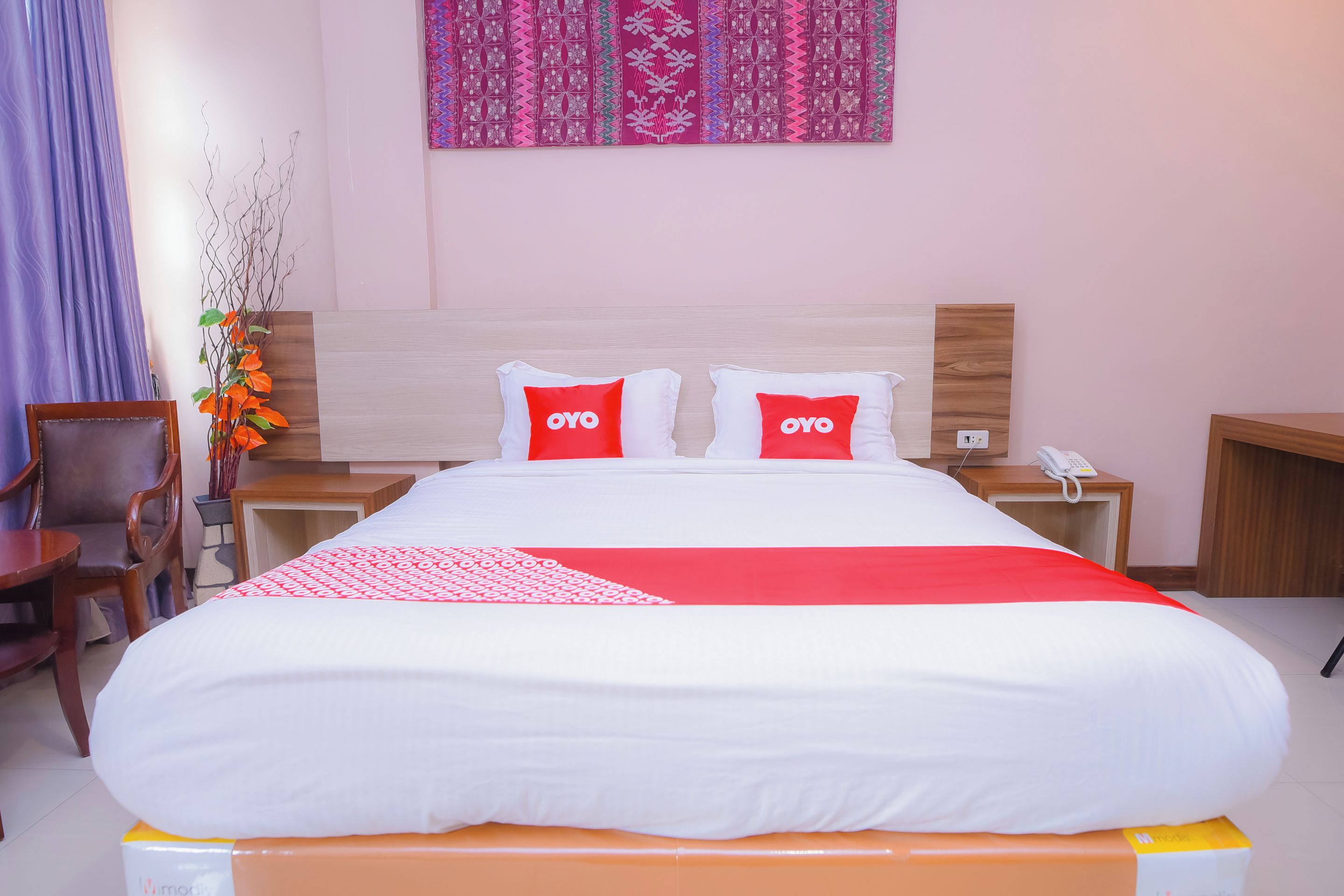 A hotel room with a bed, two pillows with 'OYO' branding, a bedside table with a phone, and a wall with patterned decor.
