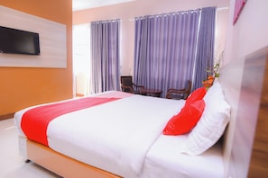 Een opgemaakt bed met een rood-witte dekbedovertrek, een flatscreen-tv aan de muur en een raam met paarse gordijnen.