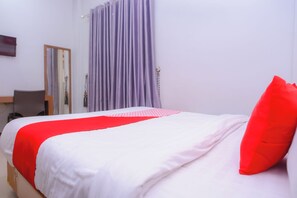 Een net opgemaakt bed met wit en rood beddengoed, een rood kussen en een paars gordijn in een eenvoudige kamer met een houten hoofdbord en een klein bureau met stoel.