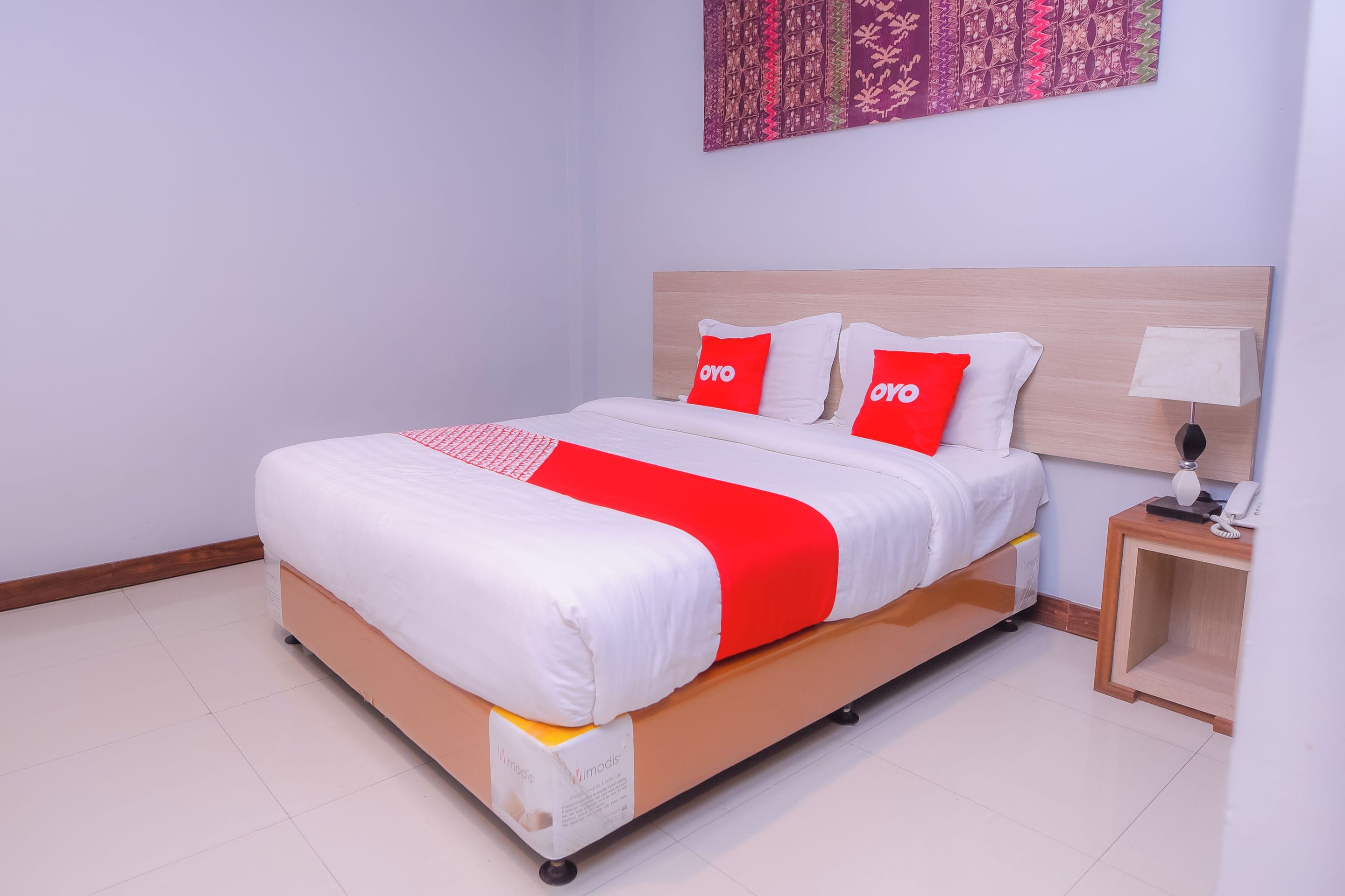 A neatly made bed with a white and red bedspread, two red pillows with "OYO" branding, a bedside table with a lamp, and a wall with a patterned tapestry.