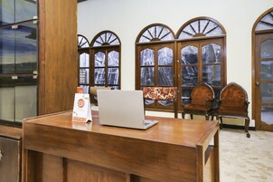 A wooden reception desk with a laptop and a sign, wooden chairs, and arched windows with a view.