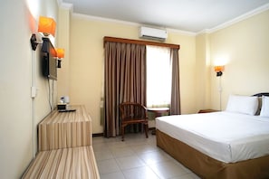 A hotel room with a bed, a desk with a lamp, a chair, and an air conditioning unit.