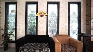 A room with a black sofa, a patterned ottoman, and a tan armchair. There are three windows with decorative glass panes and a fan hanging between them. - OYO 2015 Bandara Hotel Balikpapan (Balikpapan)