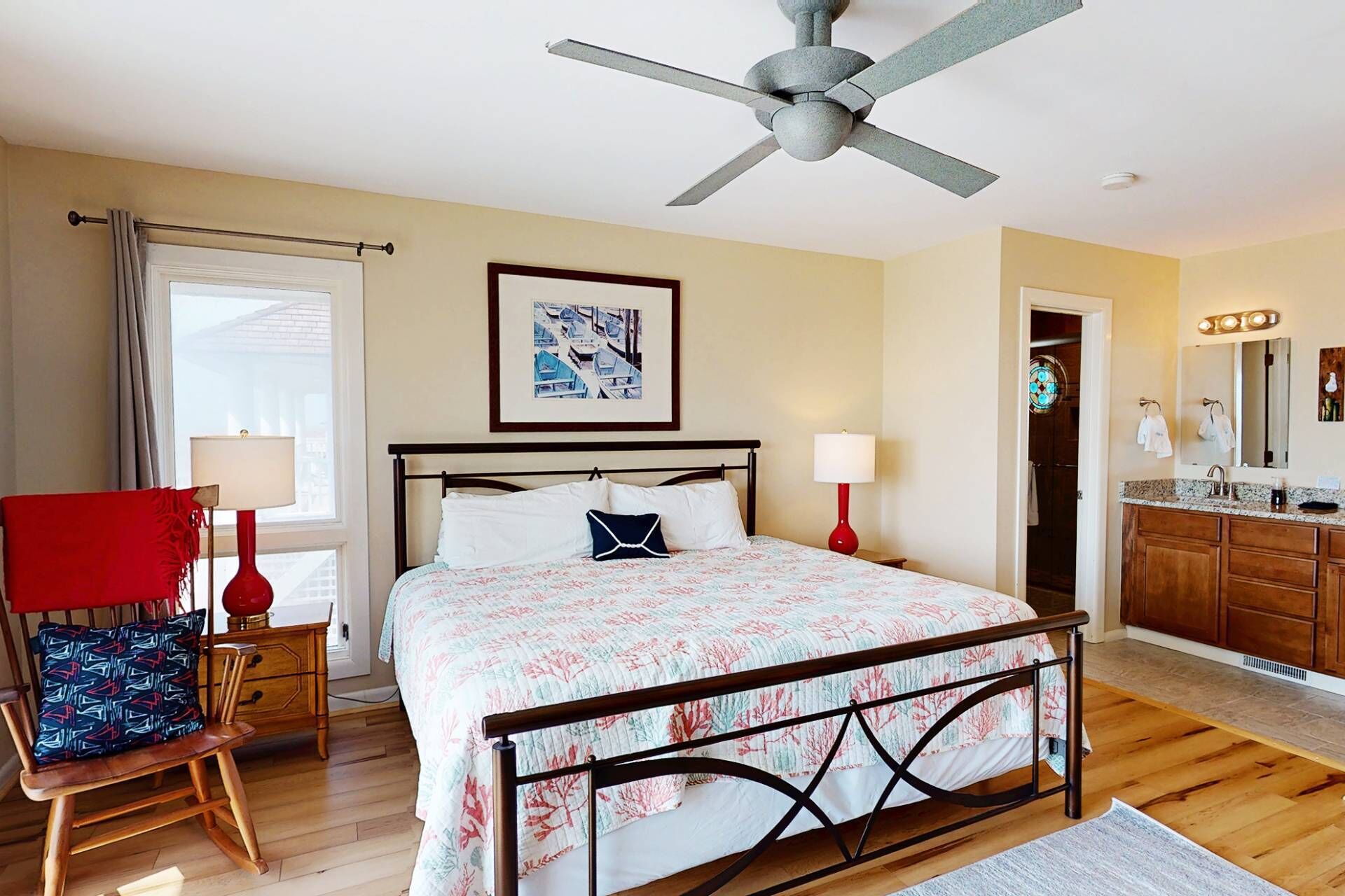 A bedroom with a bed, a ceiling fan, a window with curtains, a chair, a bedside table, a nightstand, a mirror, and a bathroom with a sink and mirror.