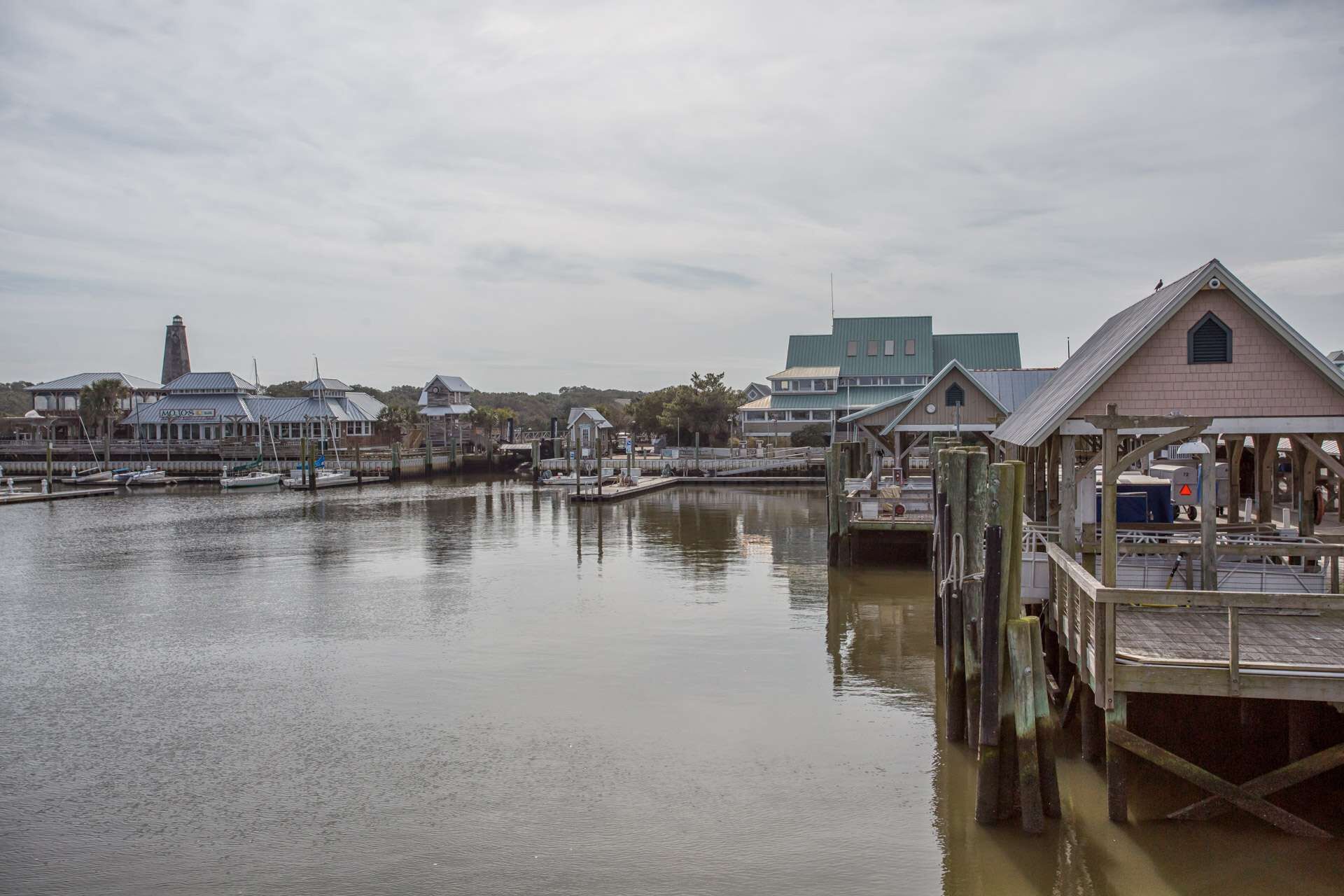 A waterfront marina with docked boats, a lighthouse, and various buildings including a restaurant and a large house.