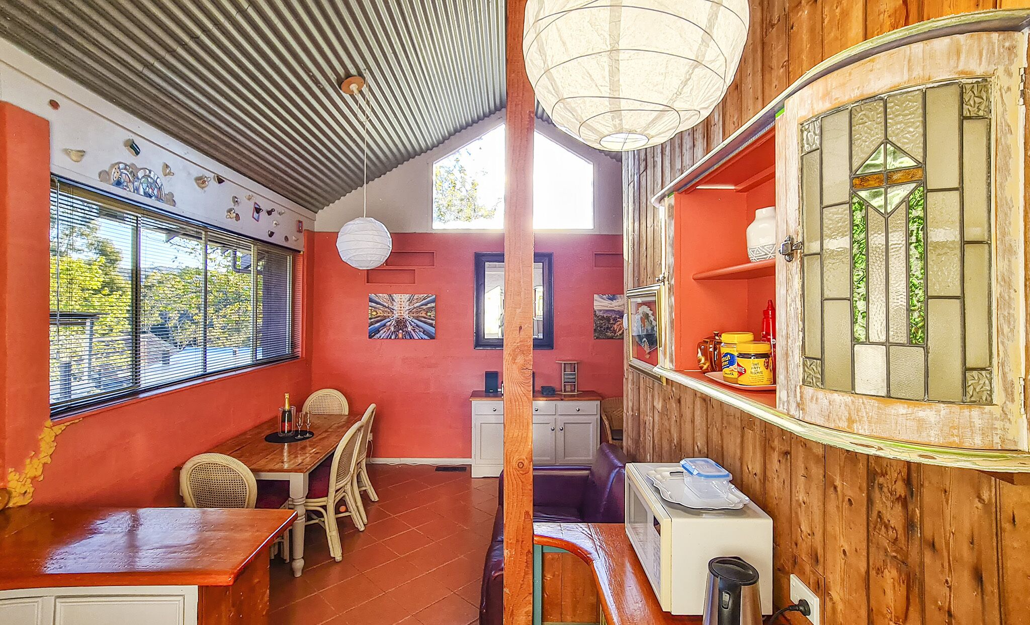 Ein Essbereich mit einem Holztisch, Stühlen und einer roten Wand. Ein Fenster mit Jalousien, eine weiße Decke und ein Holzregal mit Gläsern sind ebenfalls zu sehen.