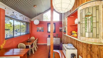 Ein Essbereich mit einem Holztisch, Stühlen und einer roten Wand. Ein Fenster mit Jalousien, eine weiße Decke und ein Holzregal mit Gläsern sind ebenfalls zu sehen.