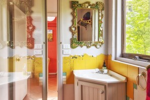 Un baño con pared de azulejos amarillos y blancos, un lavamanos blanco y un espejo decorativo con marco dorado.