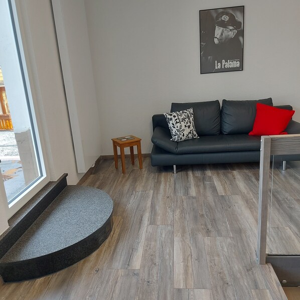 A modern living room with a dark grey sofa, a small wooden stool, and a grey pet bed. A poster of a historical figure hangs on the wall. - OK vacation apartment with separate entrance and covered terrace (Bad Lauterberg im Harz)