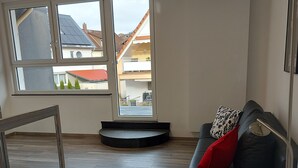 A modern living room with a black leather sofa, a glass coffee table, and a large window offering a view of neighboring houses.