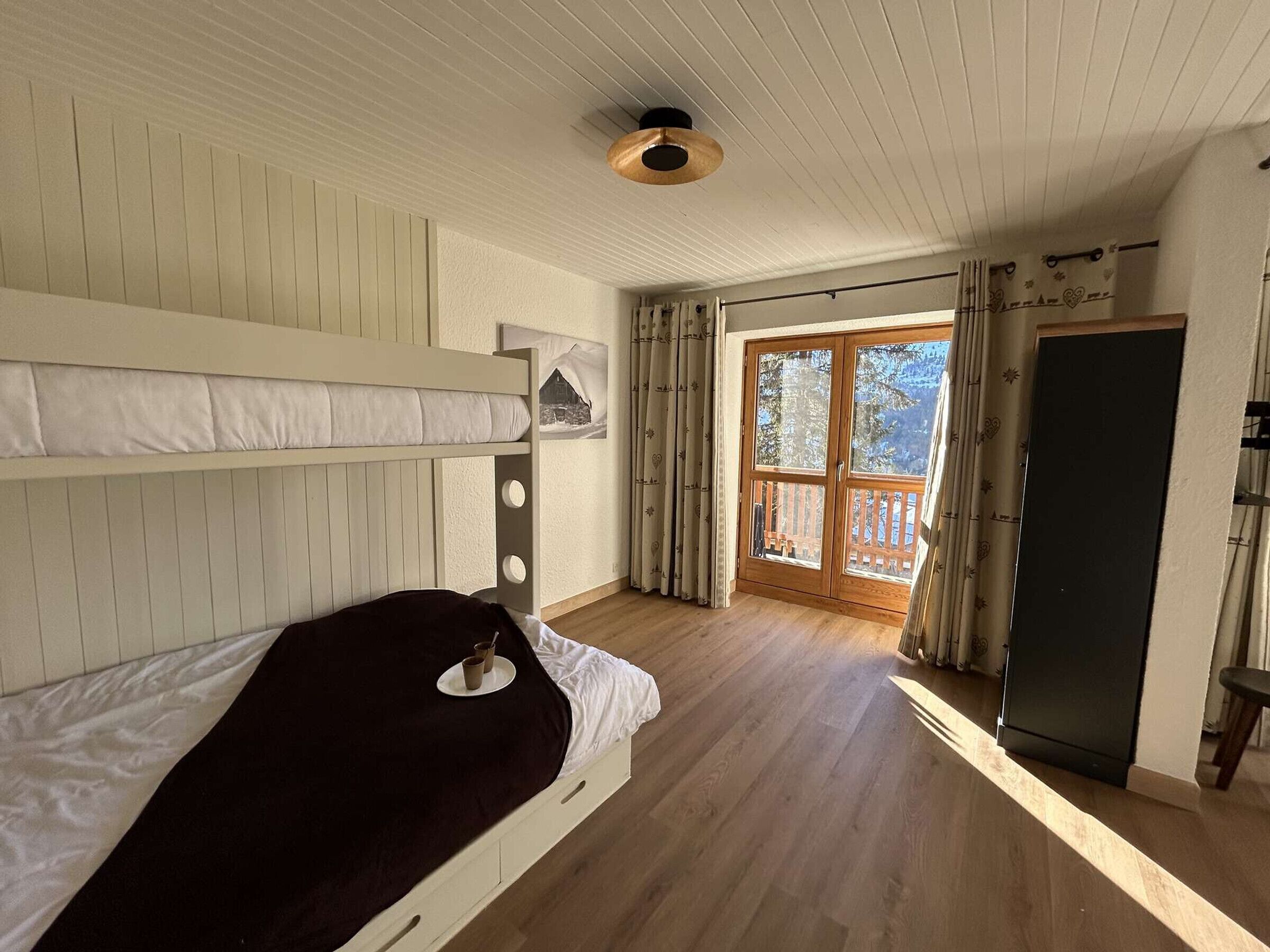 A room with a bunk bed, wooden flooring, a door leading outside, and a ceiling light fixture.