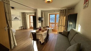Uma sala de estar acolhedora com um sofá, uma mesa de jantar posta para dois e uma grande janela com vista para uma paisagem nevada.
