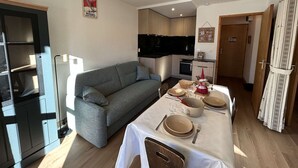 Um espaço compacto com sofá cinza, mesa de jantar posta com pratos e talheres, área de cozinha com micro-ondas e televisão, e porta de madeira.