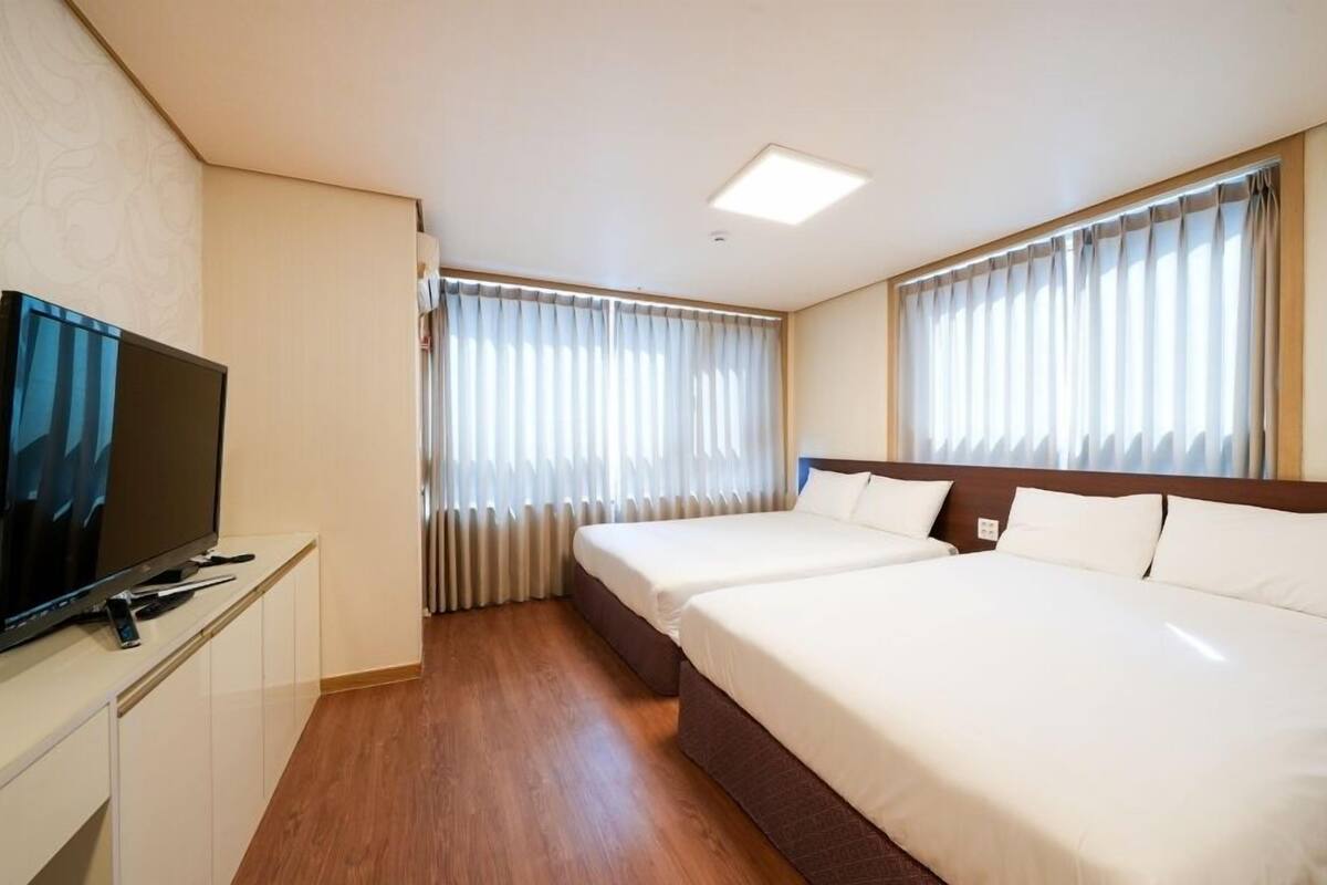 A hotel room with a TV on a desk, two beds, and large windows with curtains.
