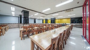 A spacious dining area with multiple wooden tables and chairs, a kitchen area with yellow walls, and a red-framed glass door. - Hotel Aventree Yeouido (Seoul)
