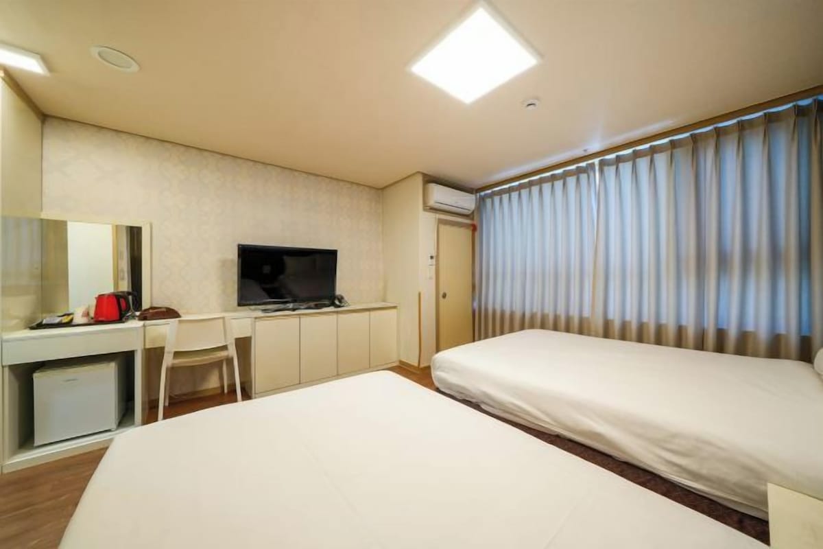 Quarto de hotel com uma cama, uma escrivaninha com televisão, uma cadeira e uma janela com cortinas.