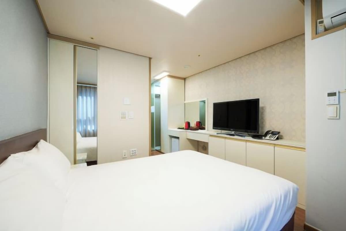 Quarto de hotel com uma cama grande, televisão em um suporte, espelho e guarda-roupa.