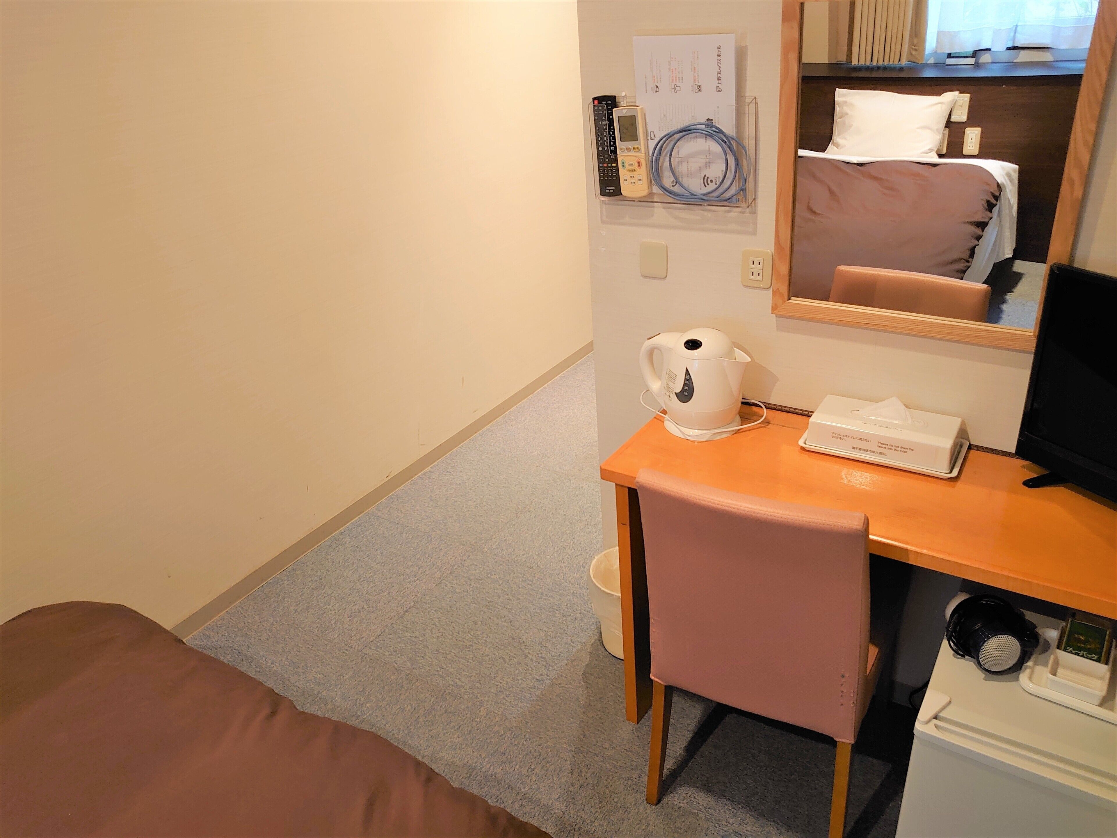A hotel room with a bed, a desk with a kettle, a remote control, and a monitor.