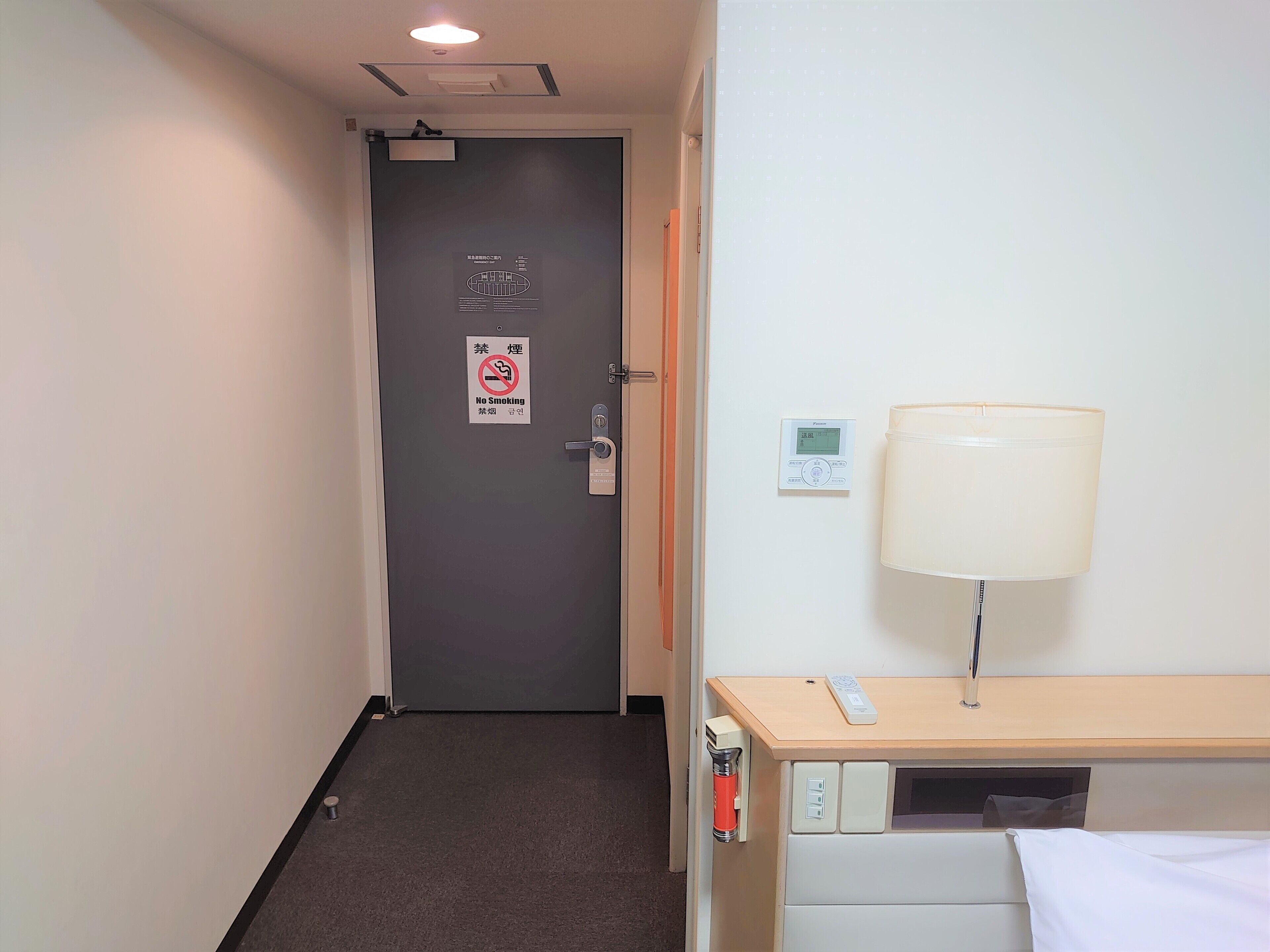A hotel room with a door, a no-smoking sign, a thermostat, a lamp, a bedside table, and a bed.