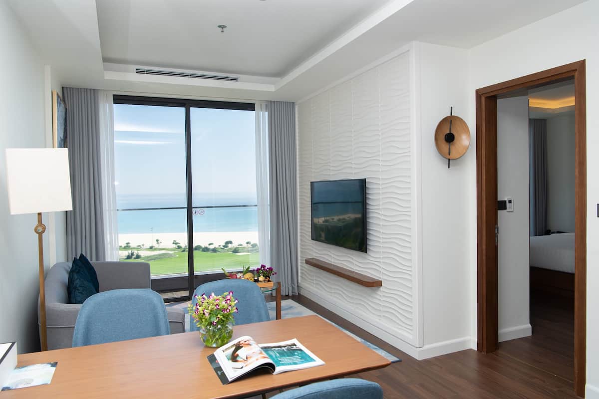 Ein Hotelzimmer mit großem Fenster und Meerblick, einem an der Wand befestigten Flachbildfernseher, einem Holztisch mit einer Zeitschrift und einem blauen Sessel.