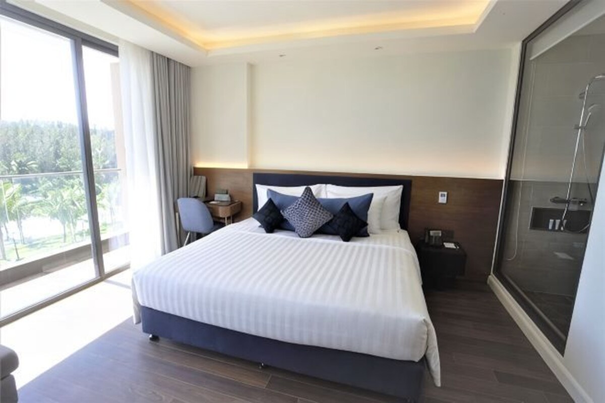 Une chambre d’hôtel avec un grand lit, un bureau, une chaise, une grande fenêtre et une porte coulissante.