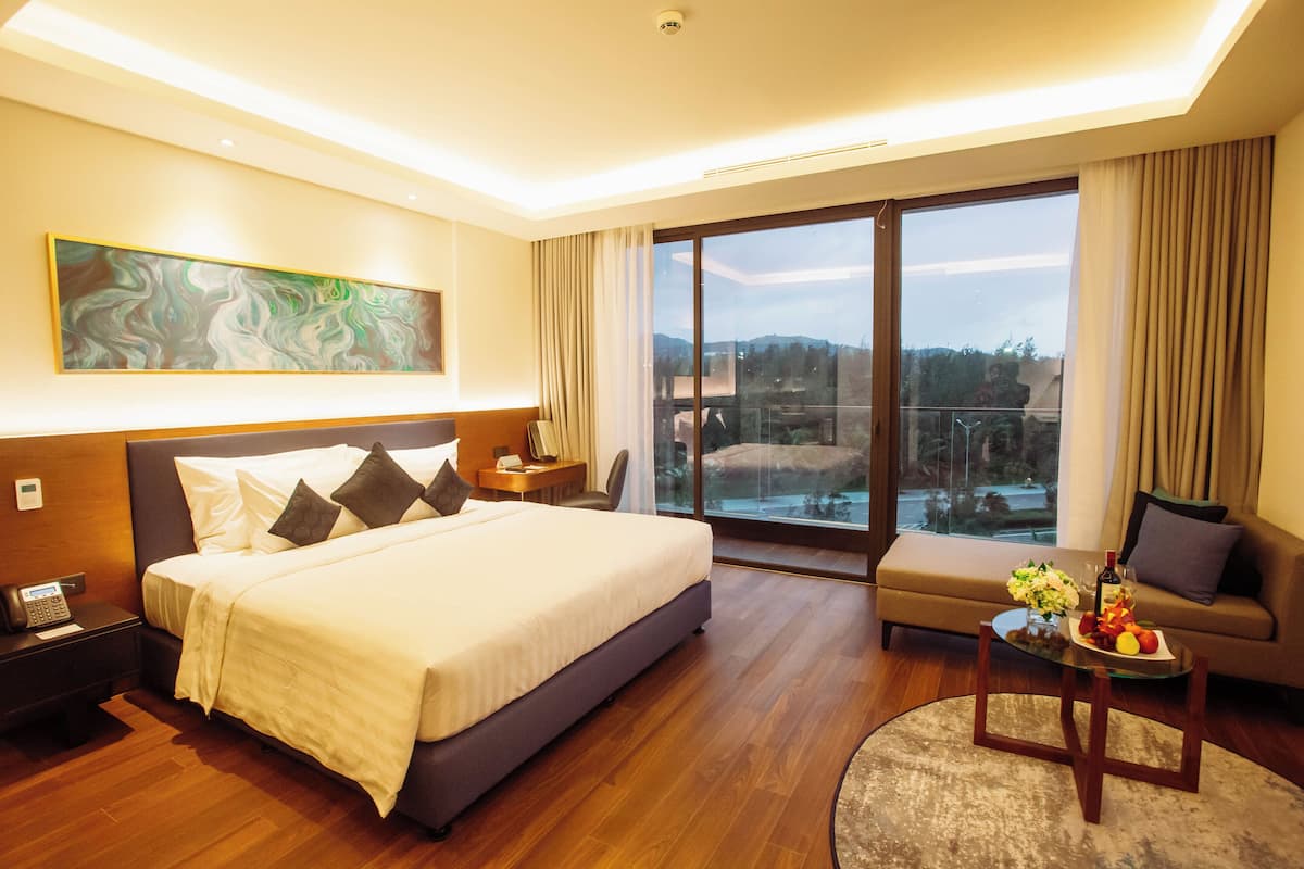 Ein modernes Hotelzimmer mit einem großen Bett, einer Couch, einem Esstisch mit Obst und Blumen, einem Bild an der Wand und einem Balkon mit Blick.