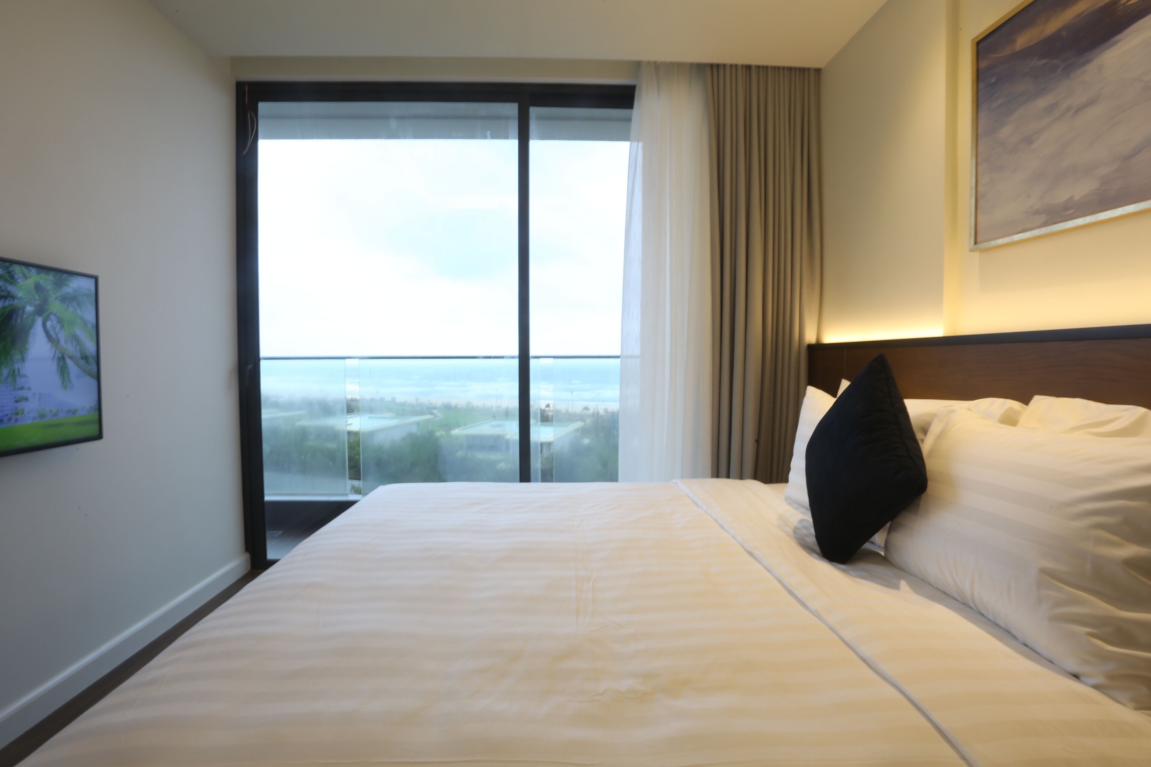 A hotel room with a large bed, a view through a glass door, and a painting on the wall.