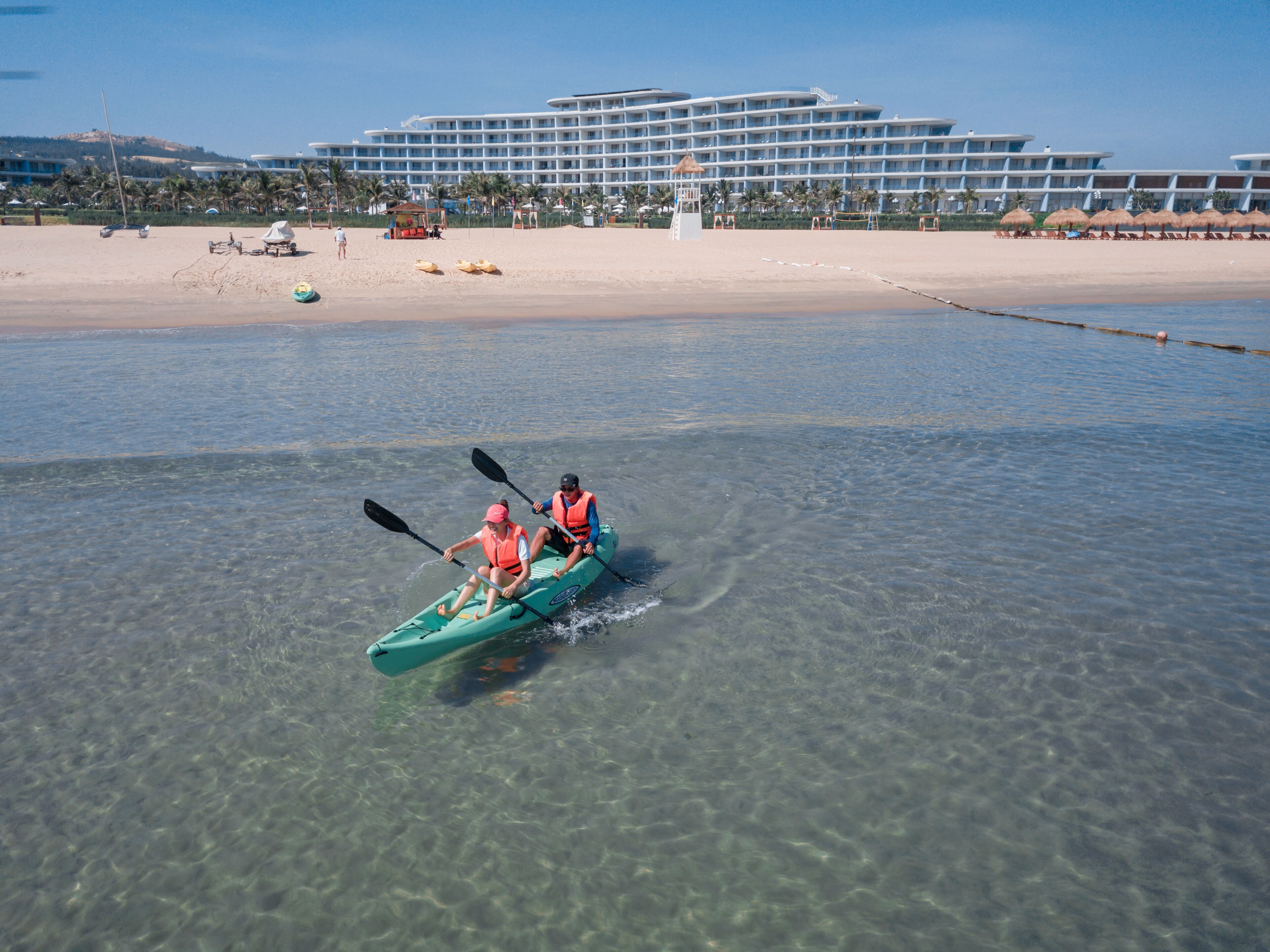 Un couple fait du kayak sur une plage de sable fin, avec un grand hôtel en arrière-plan.