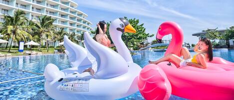 Una piscina con dos grandes flotadores de cisne hinchables, uno blanco y otro rosa, y un edificio moderno de hotel al fondo.