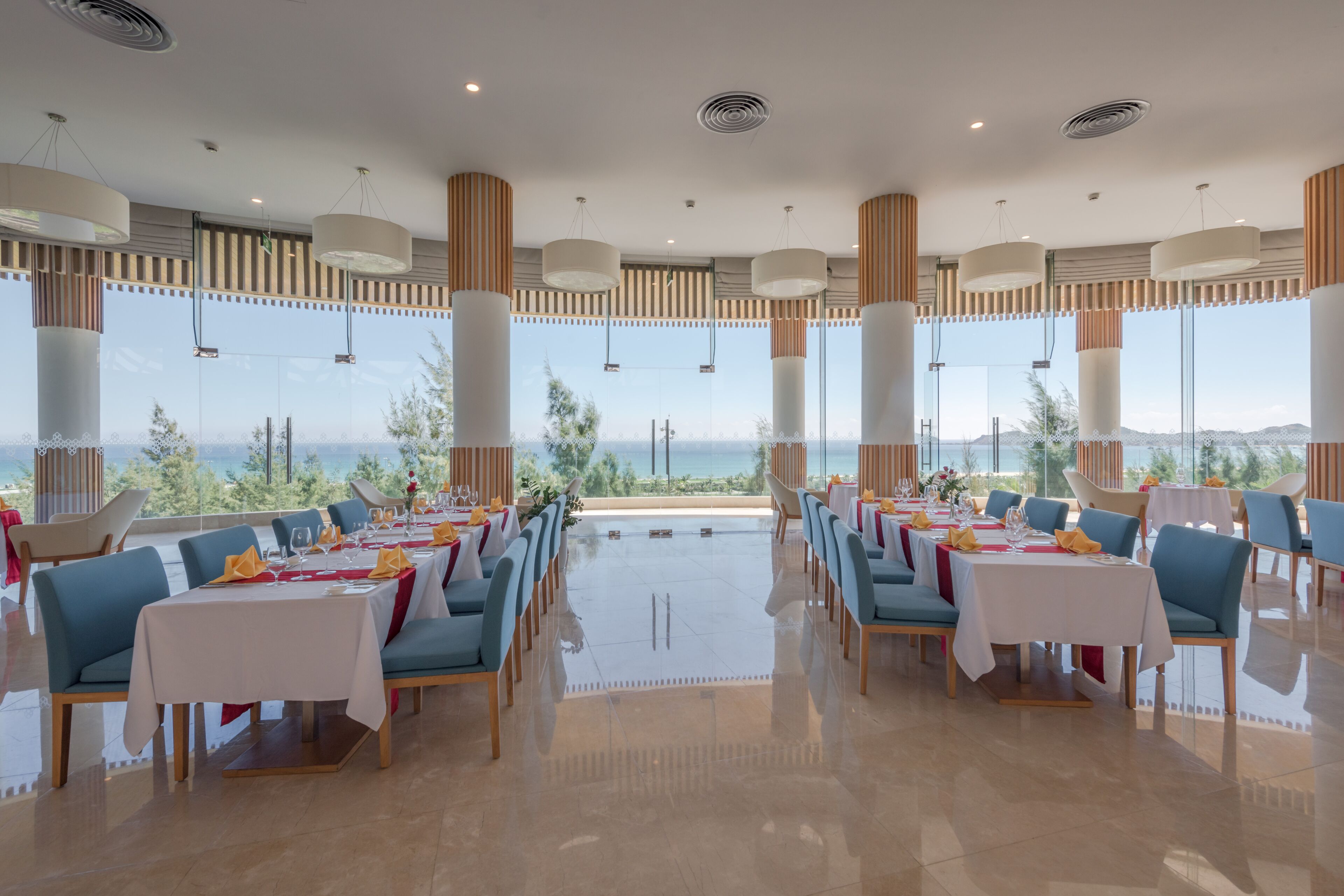 A restaurant with tables set for dining, large windows with a view of the sea, and modern pendant lighting.