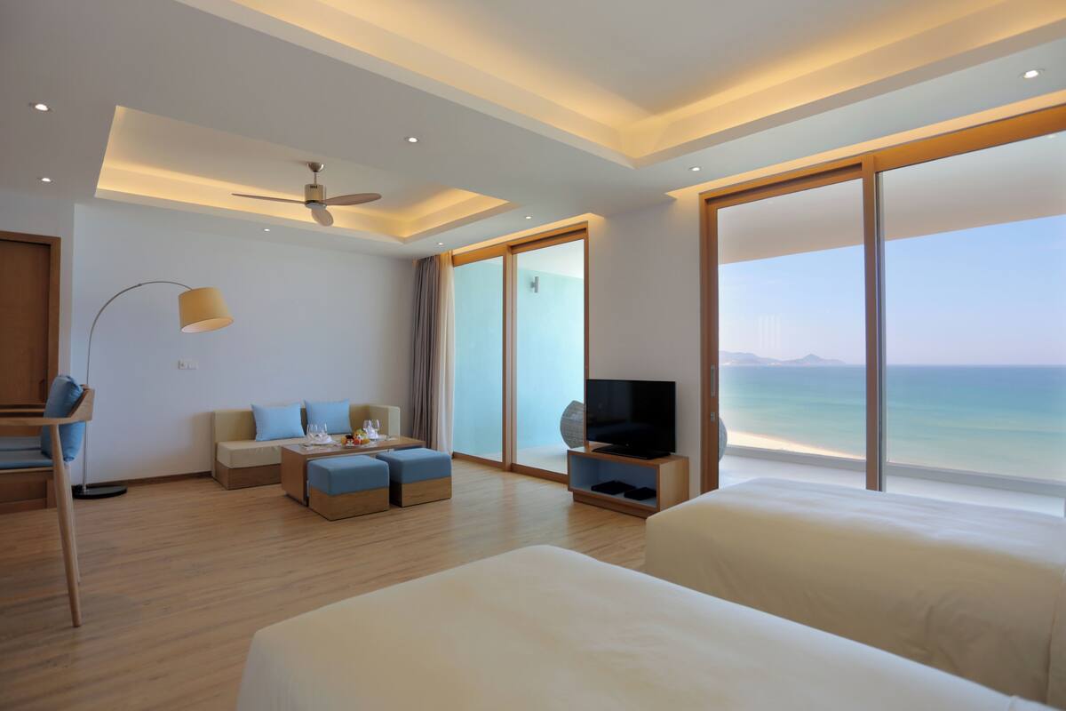 Ein modernes Hotelzimmer mit einem großen Bett, einer Sitzecke mit Fernseher und einem Balkon mit Meerblick.