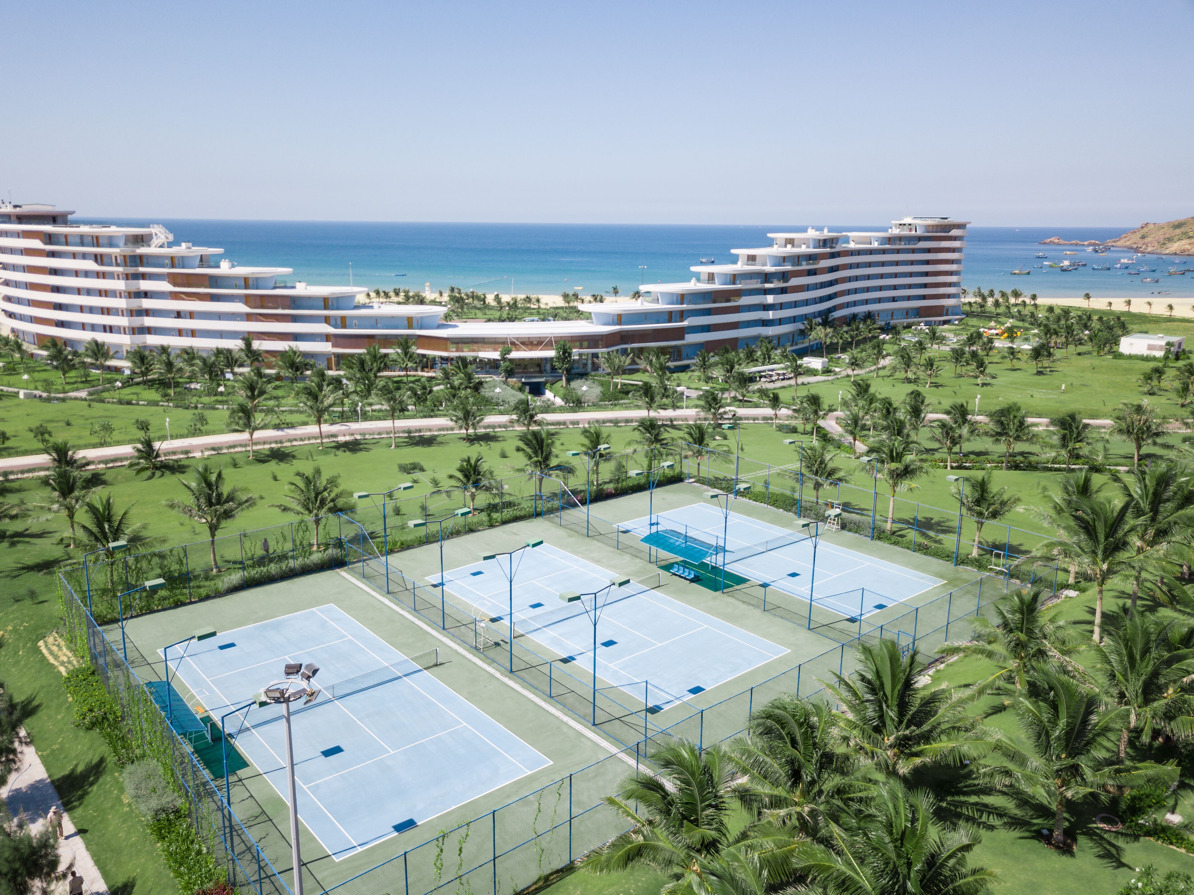 Ein Resort mit mehreren Tennisplätzen, umgeben von Palmen, und ein großes Gebäude mit Pool im Hintergrund.