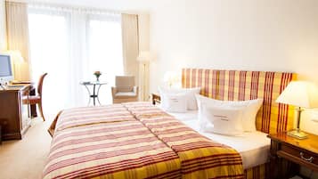 A hotel room with a large bed, a desk with a computer, a chair, a small table with a vase, and a window with curtains.