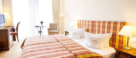 Habitación de hotel con una cama grande, un escritorio con computadora, una silla, una mesita con un jarrón y una ventana con cortinas.