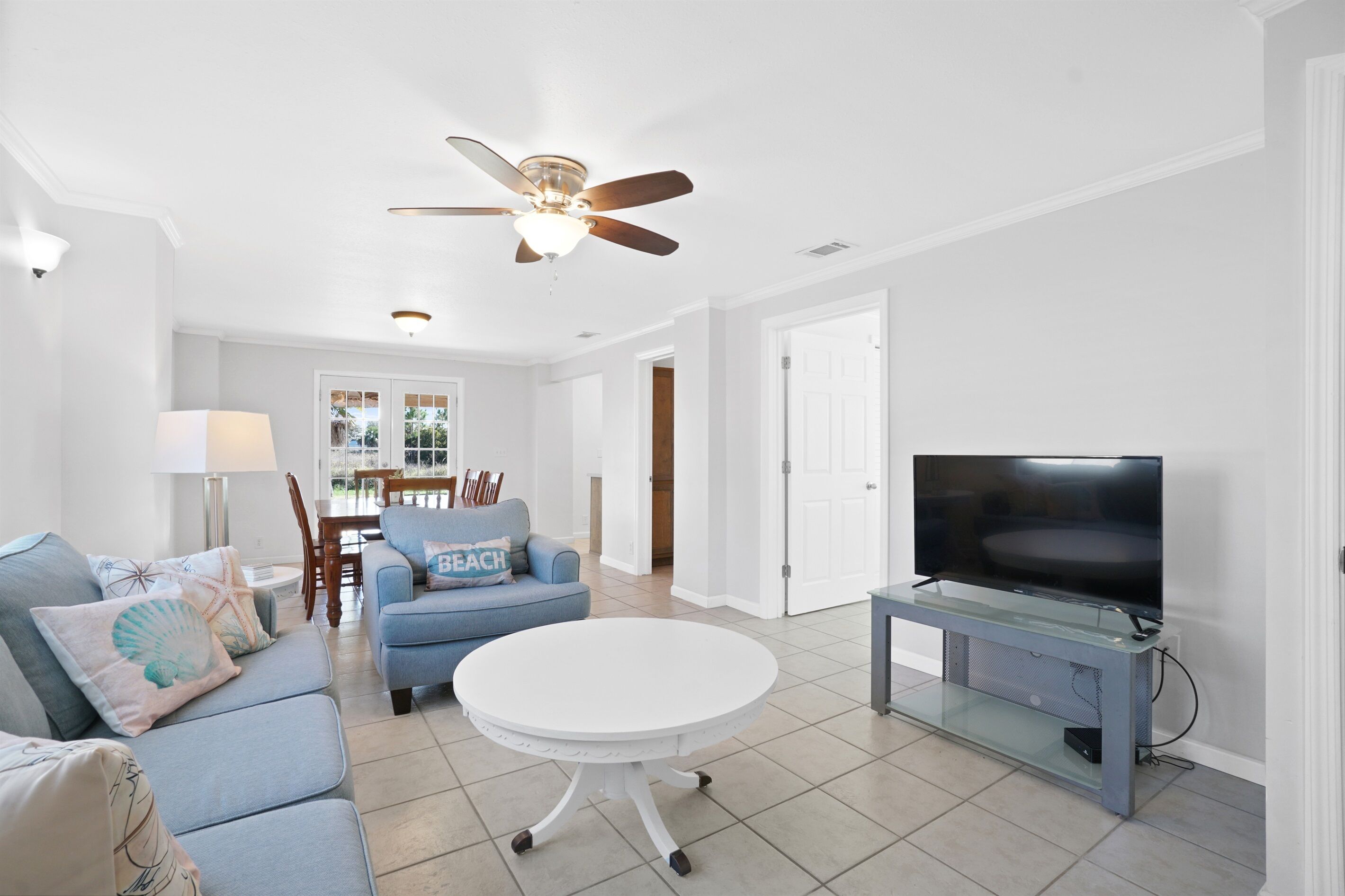 A living room with a blue sofa, a round white table, a TV on a glass stand, a ceiling fan, and a door leading to another room.