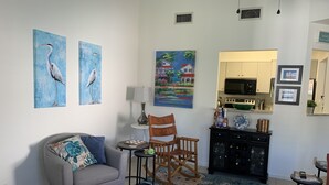 A living room with a grey armchair, a wooden rocking chair, a patterned rug, a small side table, and wall art including two paintings of birds and a landscape. - St Simons Island,  2 bedroom, 2 bath condo, short walk to village and beaches. (St. Simons)