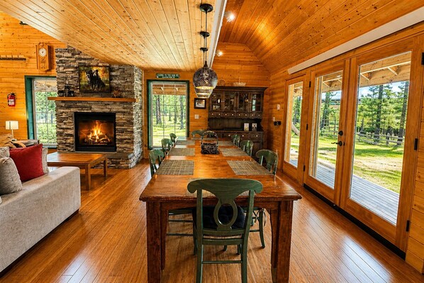 A wooden interior with a fireplace, dining table, chairs, and large windows.