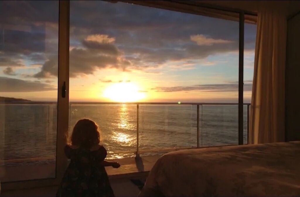 Un enfant observe le coucher de soleil sur l’océan depuis un balcon.