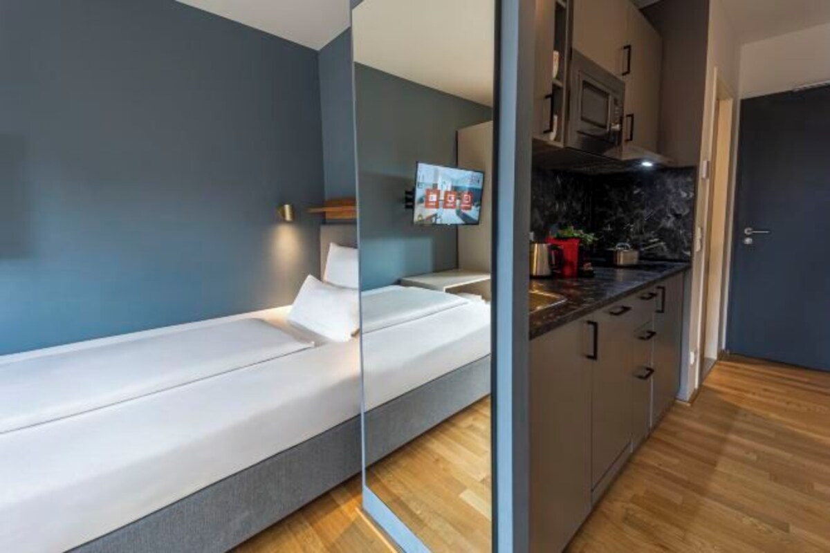 Een moderne kamer met een bed, een televisie, een keukenhoek met kastjes en een magnetron, en een deur die naar een andere kamer leidt.