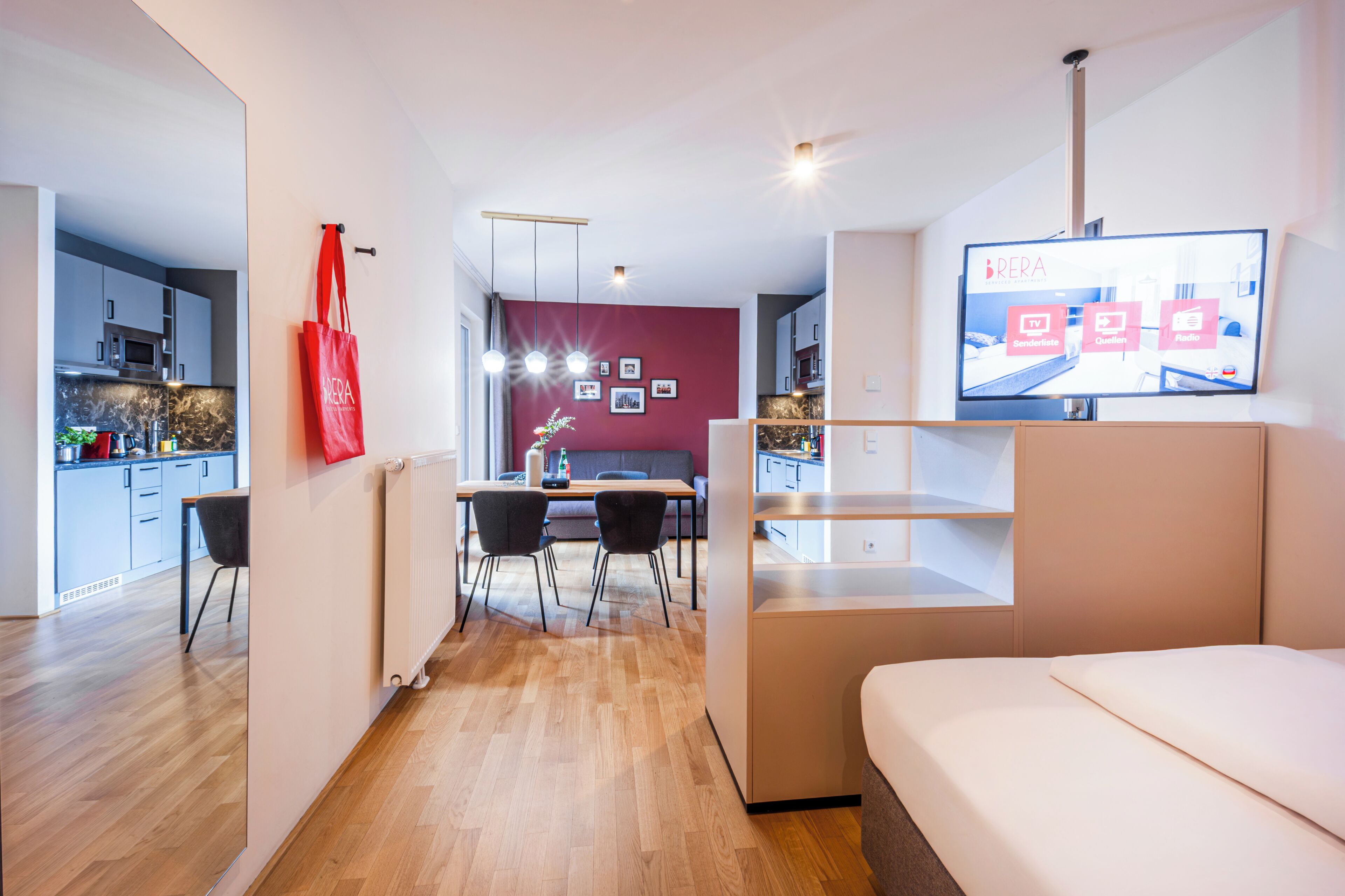 A modern hotel room with a bed, a TV, a dining area with chairs, and a kitchen in the background.