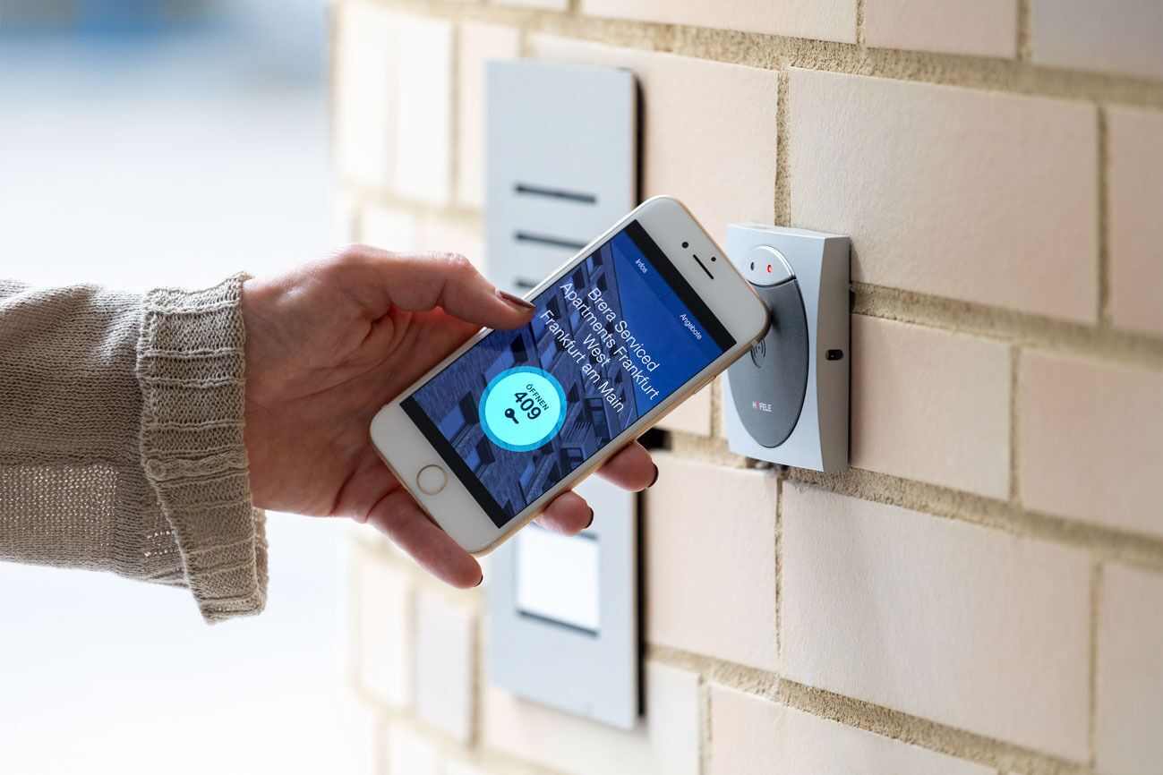 A hand holding a smartphone displaying a digital interface with a circular icon and text, positioned near a wall-mounted device with a rectangular display.