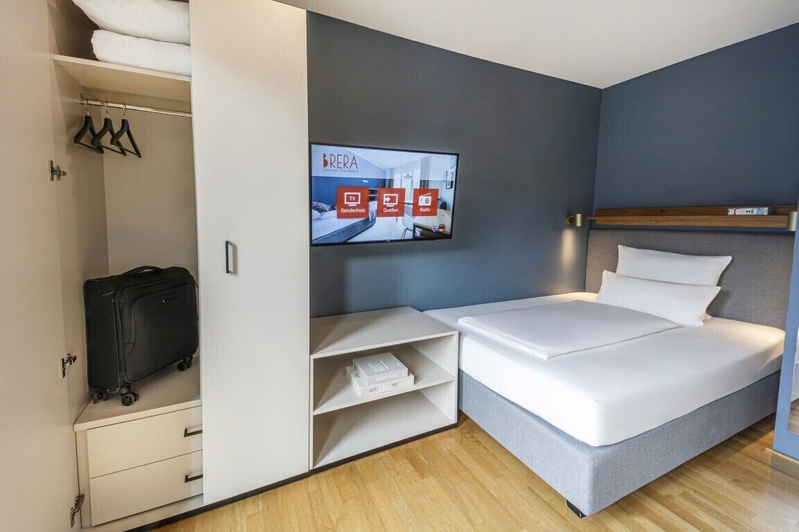 Habitación de hotel con cama, televisor en la pared, armario y maleta.